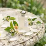 Elegant glass perfume bottle with silver cap and tassel, set among jasmine blossoms on natural stone for a luxury beauty vibe.