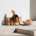 Alwa perfume bottle with gold cap and amber liquid, placed on wooden blocks with a bowl of sliced pe.