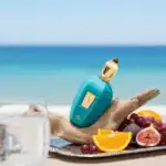 Fendi Pura perfume bottle with a turquoise finish and gold cap, set against a beach backdrop with fr.
