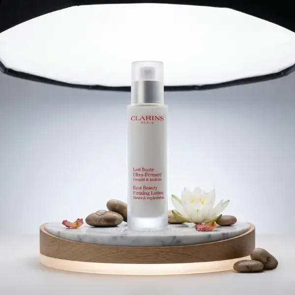 Clarins Bust Beauty Firming Lotion in a sleek white bottle with a pump, displayed on a marble and wo.