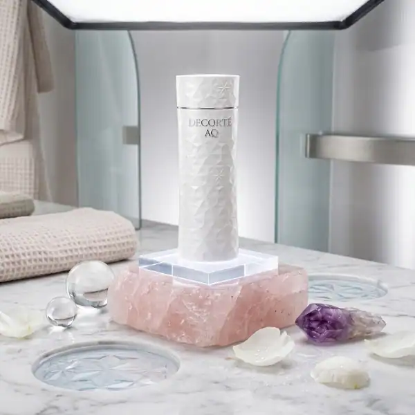 Elegant Decorté AQ skincare bottle displayed on pink crystal with decorative spheres and petals.