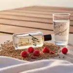 Aqua perfume bottle with raspberries and grains, next to a glass of sparkling water on a wooden surf.