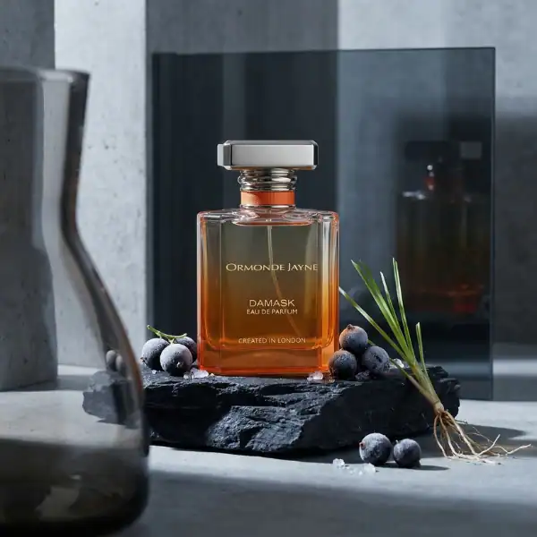 Ormonde Jayne Damask fragrance bottle with blueberries and green leaves, on a dark stone surface.